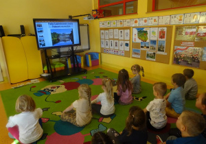Grupa dzieci siedzi przed ekranem mobilnym, oglądają prezentację multimedialną na temat Warszawy.